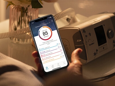 Someone sitting on a bed, holding a mobile phone with the ResMed myAir app on it in one hand and a CPAP mask in the other, with a CPAP device on the bedside table.